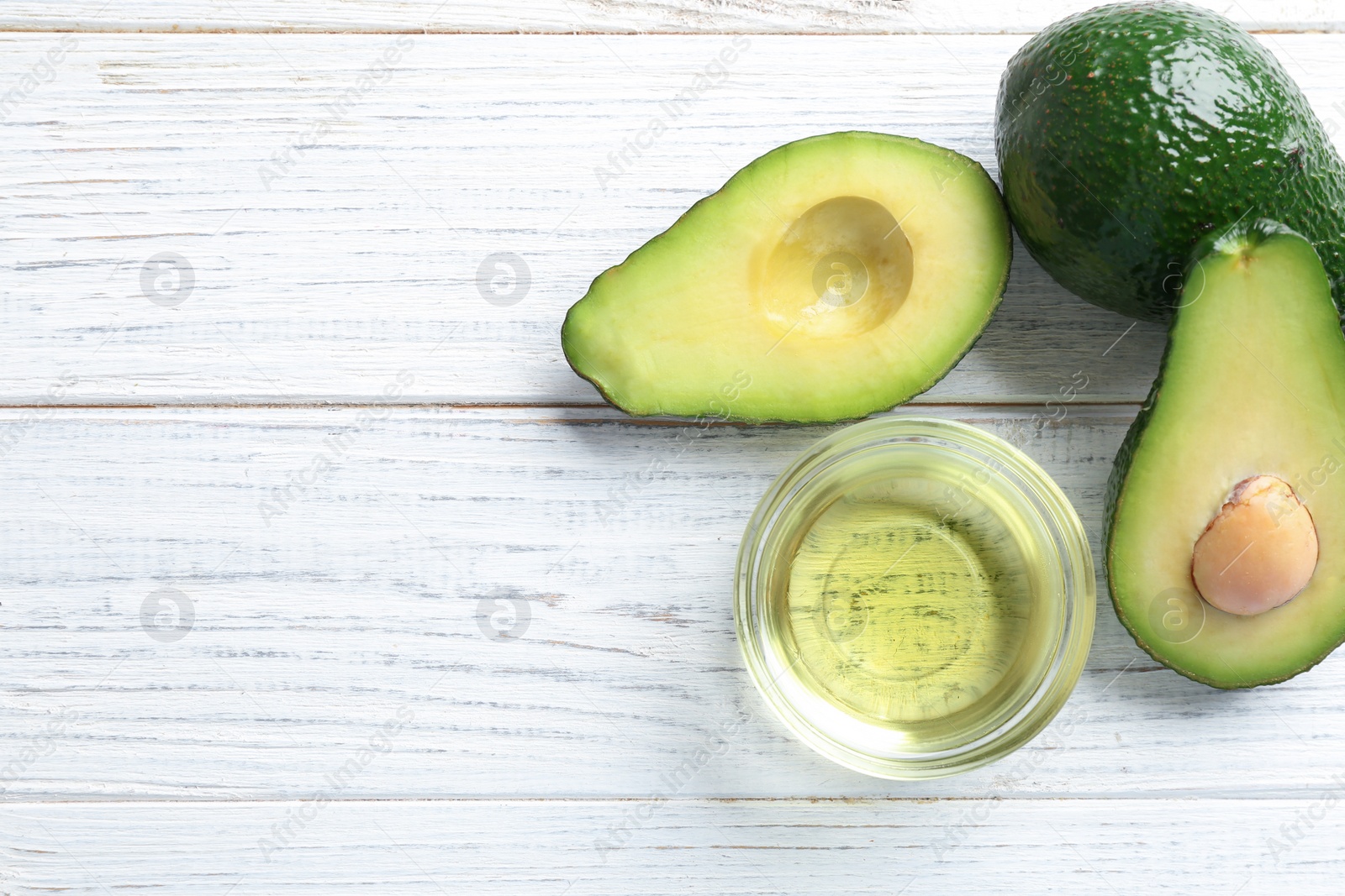 Photo of Bowl of natural oil and avocados on white wooden background, flat lay. Space for text