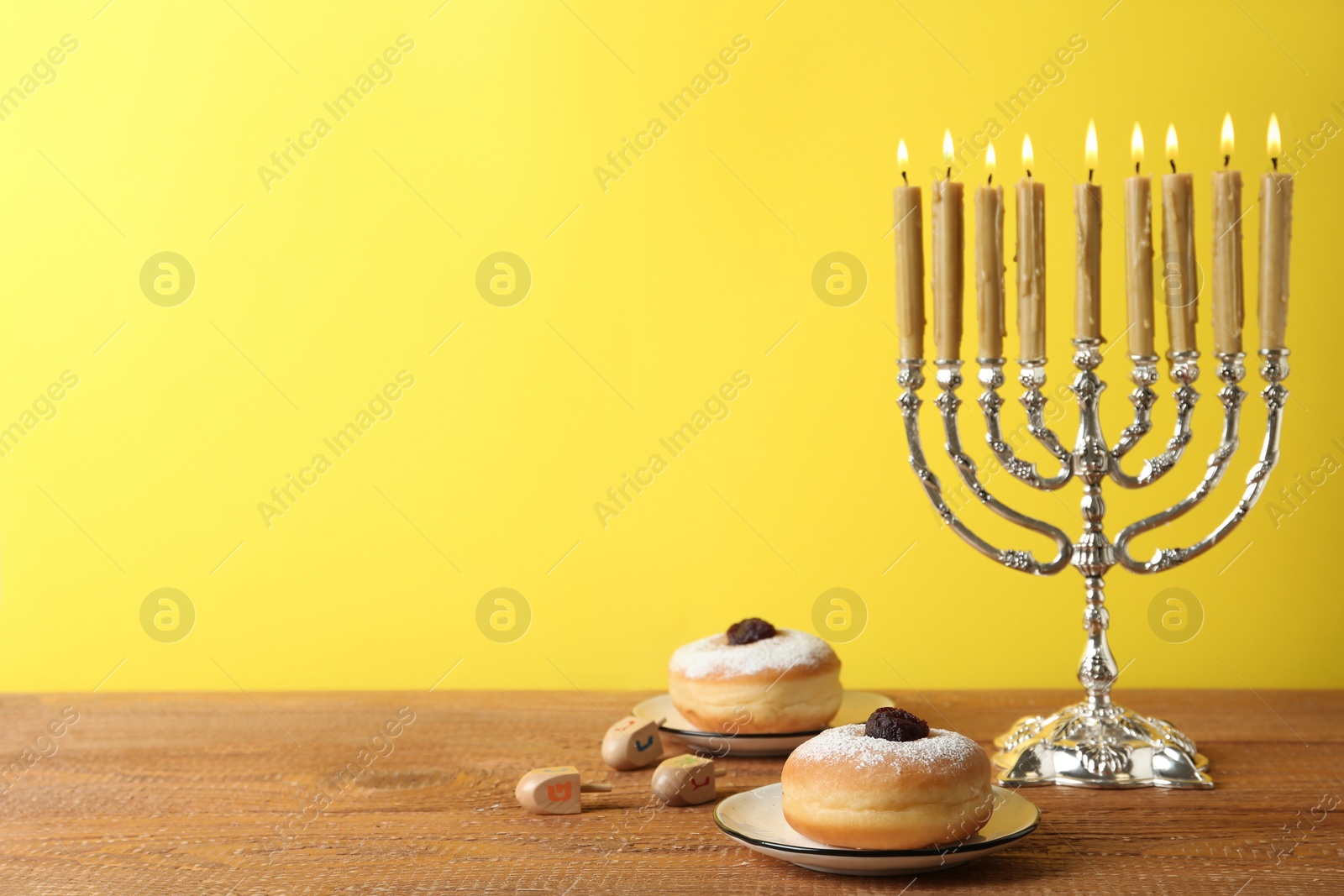 Photo of Silver menorah, dreidels with He, Pe, Nun, Gimel letters and sufganiyot on wooden table against yellow background, space for text. Hanukkah symbols
