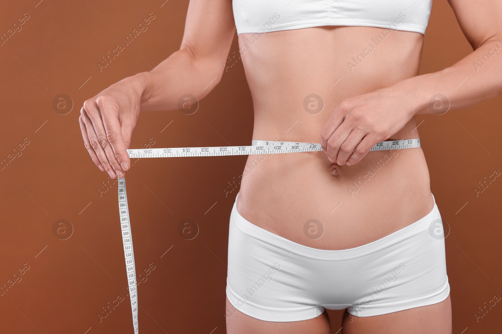 Photo of Slim woman measuring waist with tape on brown background, closeup. Weight loss