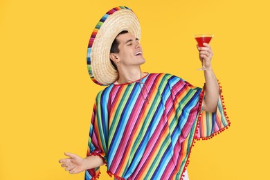 Photo of Young man in Mexican sombrero hat and poncho with cocktail on yellow background