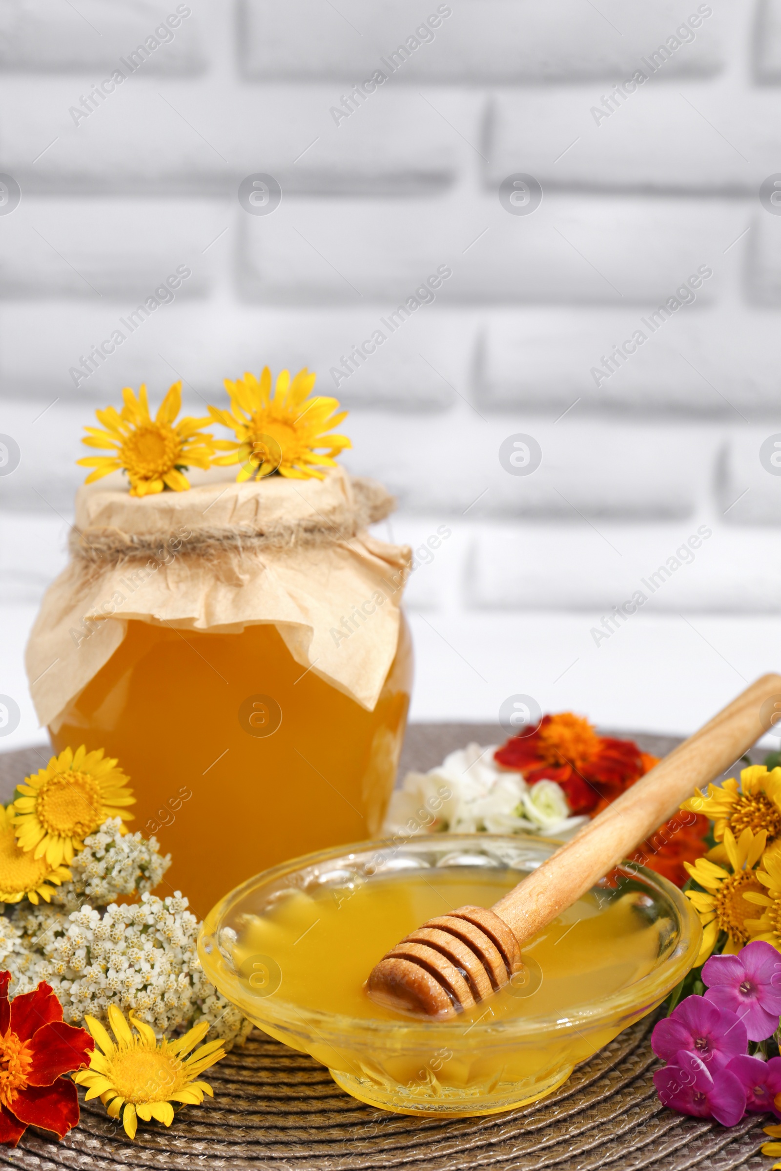 Photo of Delicious honey with dipper and different flowers on wicker mat against white brick wall