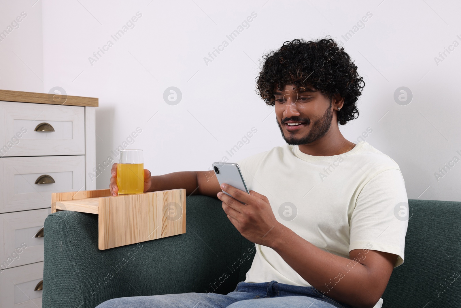 Photo of Happy man using smartphone and holding glass of juice on sofa with wooden armrest table at home