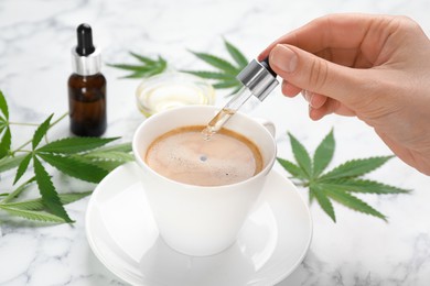 Photo of Woman dripping THC tincture or CBD oil into cup of coffee at white marble table, closeup