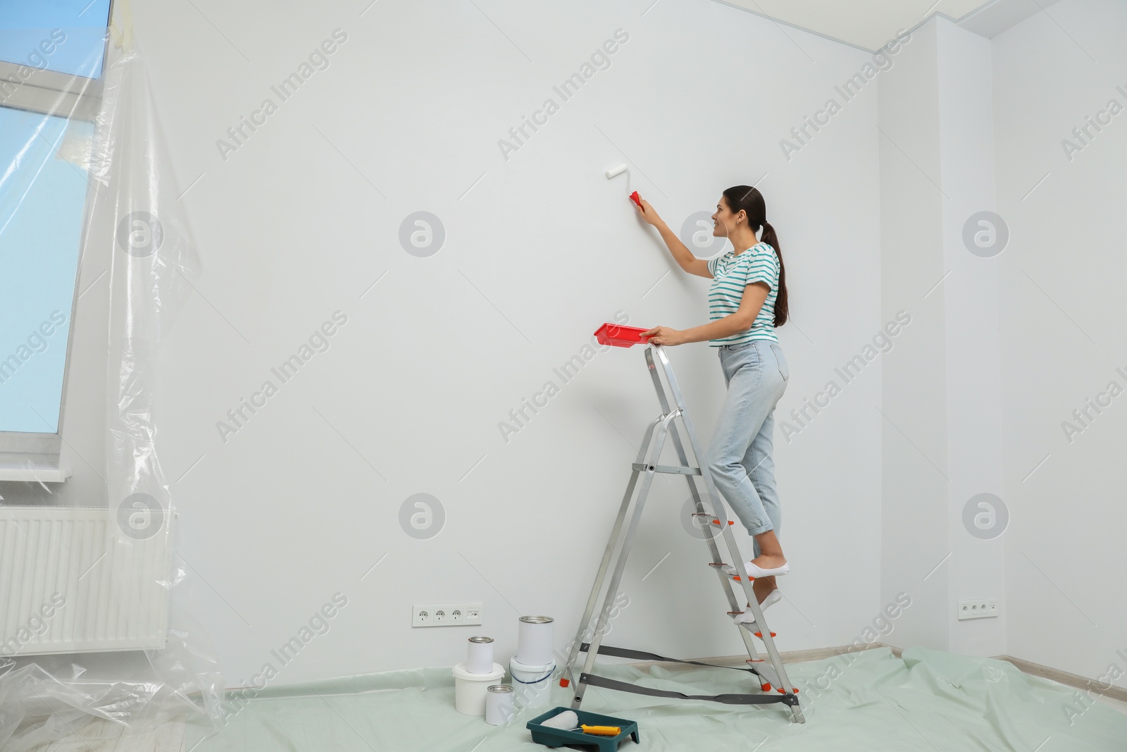 Photo of Woman standing on metallic folding ladder and painting wall indoors, space for text