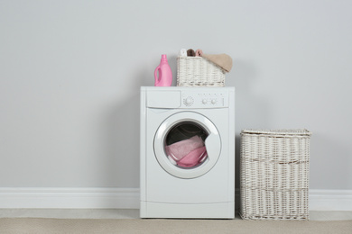 Modern washing machine with laundry, wicker baskets and detergent near white wall