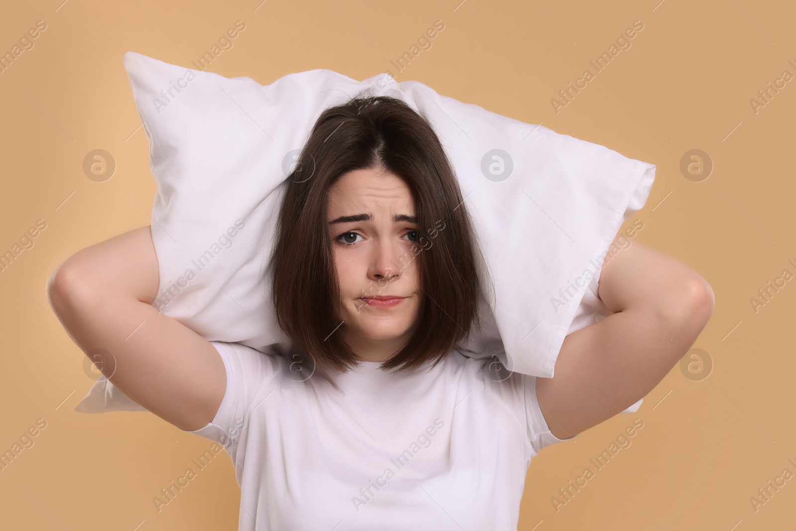 Photo of Unhappy young woman covering ears with pillow on beige background. Insomnia problem