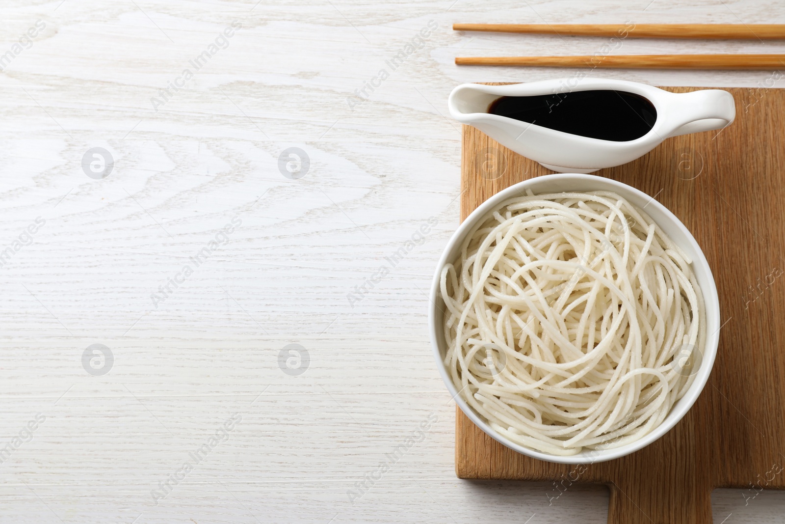 Photo of Bowl of rice cooked noodles and soy sauce served on white wooden table, flat lay. Space for text