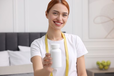 Photo of Happy young woman with bottle of pills and measuring tape in room. Weight loss