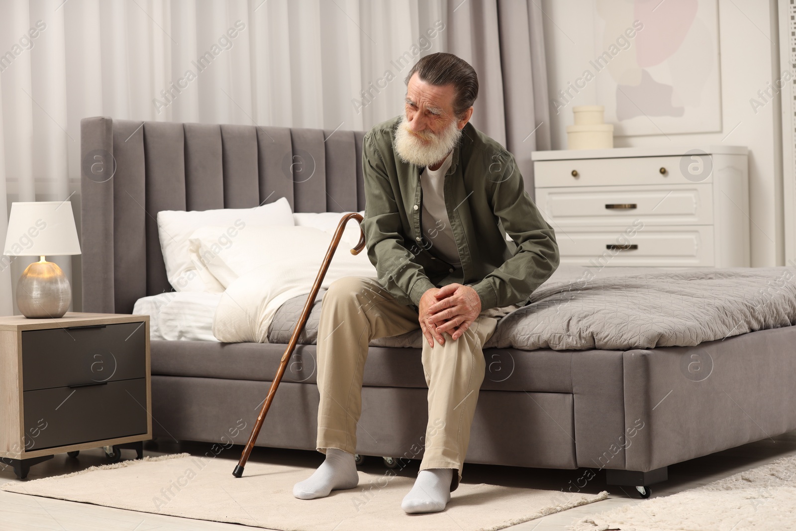 Photo of Senior man with walking cane suffering from knee pain on bed at home. Rheumatism symptom