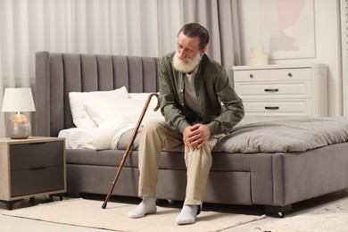 Senior man with walking cane suffering from knee pain on bed at home. Rheumatism symptom