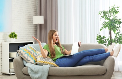 Photo of Emotional young woman with laptop celebrating victory on sofa at home