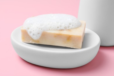 Photo of Dish with soap bar and fluffy foam on pink background, closeup
