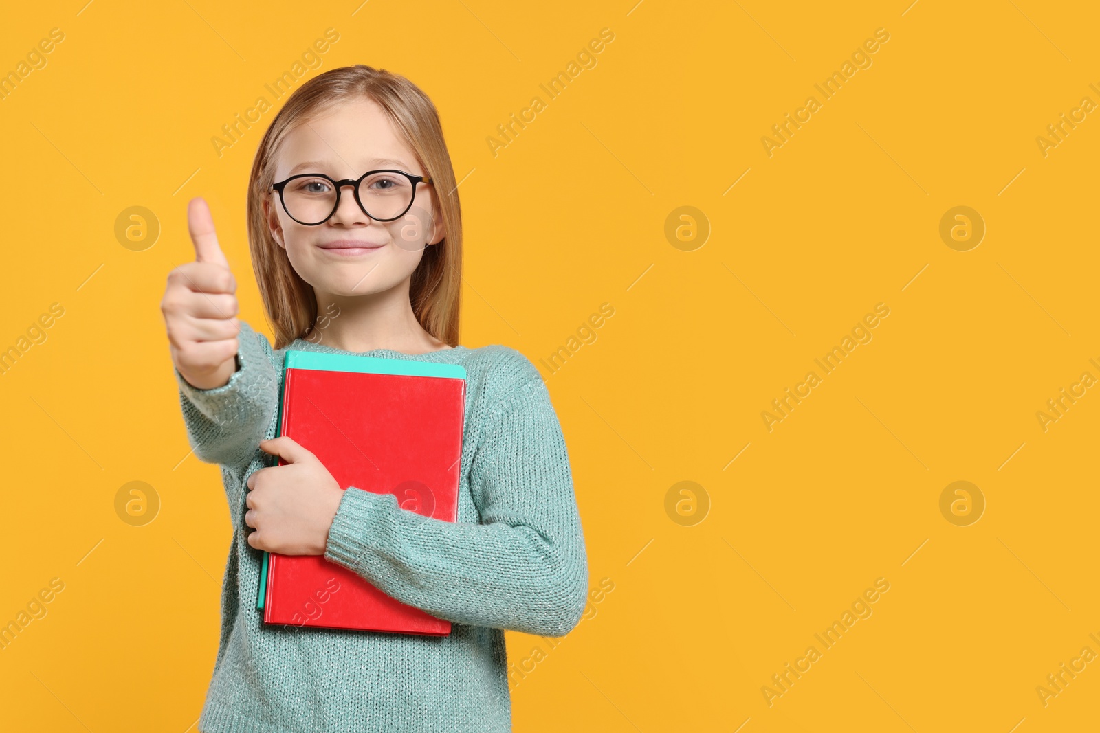 Photo of Portrait of cute girl in glasses with books showing thumb up on orange background. Space for text