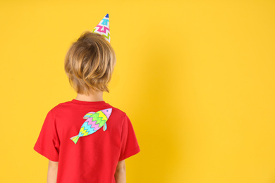 Photo of Little boy with paper fish on back against yellow background, space for text. April fool's day