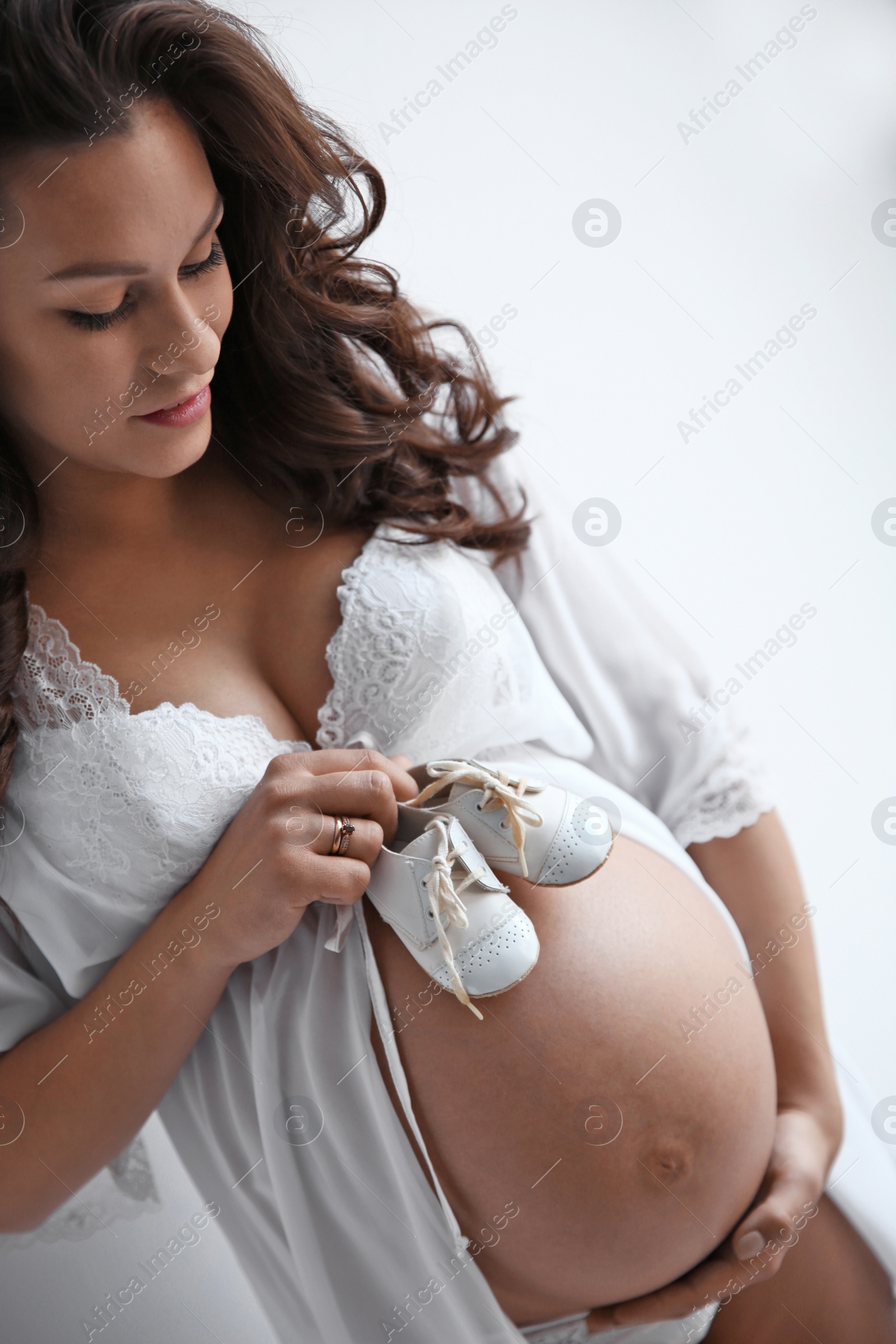 Photo of Young pregnant woman in lace nightgown holding baby shoes on light background