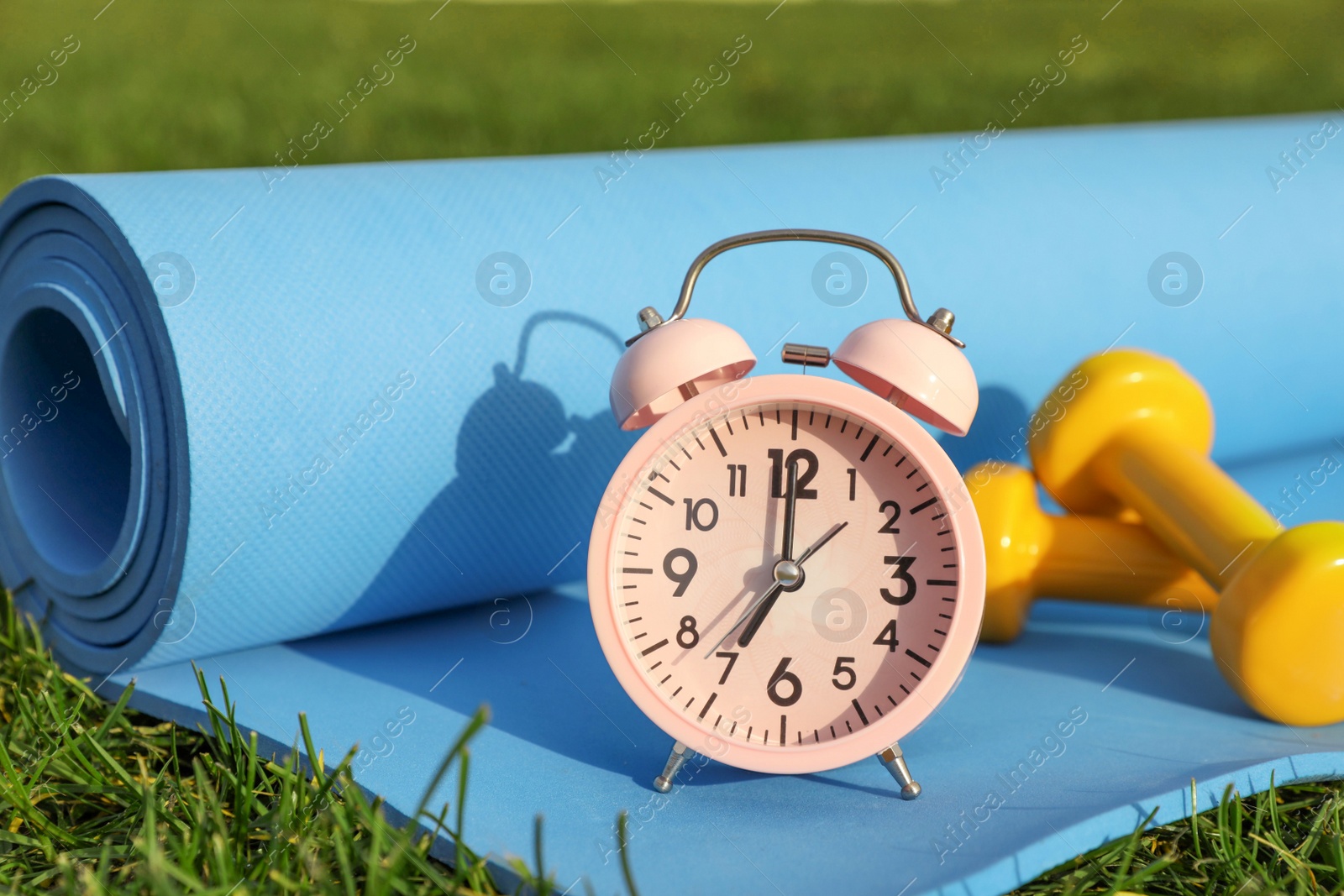 Photo of Alarm clock, dumbbells and fitness mat on green grass outdoors. Morning exercise