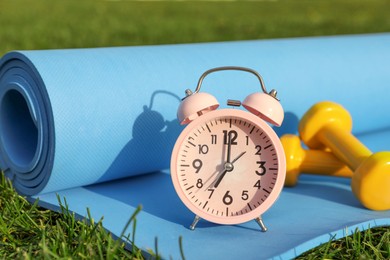 Photo of Alarm clock, dumbbells and fitness mat on green grass outdoors. Morning exercise