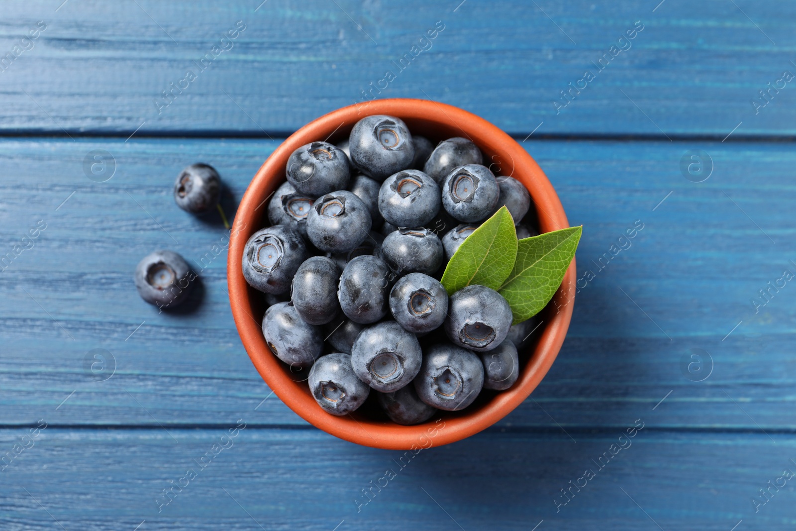 Photo of Tasty fresh blueberries with green leaves in bowl on blue wooden table, flat lay