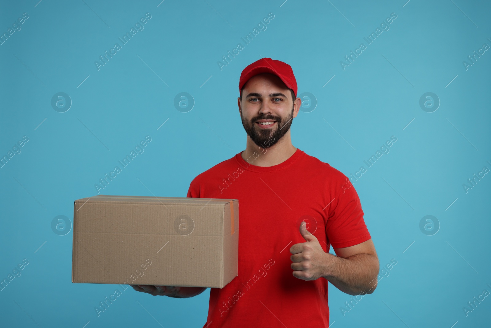 Photo of Happy young courier with parcel showing thumb up on light blue background