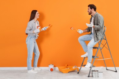 Photo of Happy woman and man with painting equipment near freshly painted orange wall indoors. Interior design