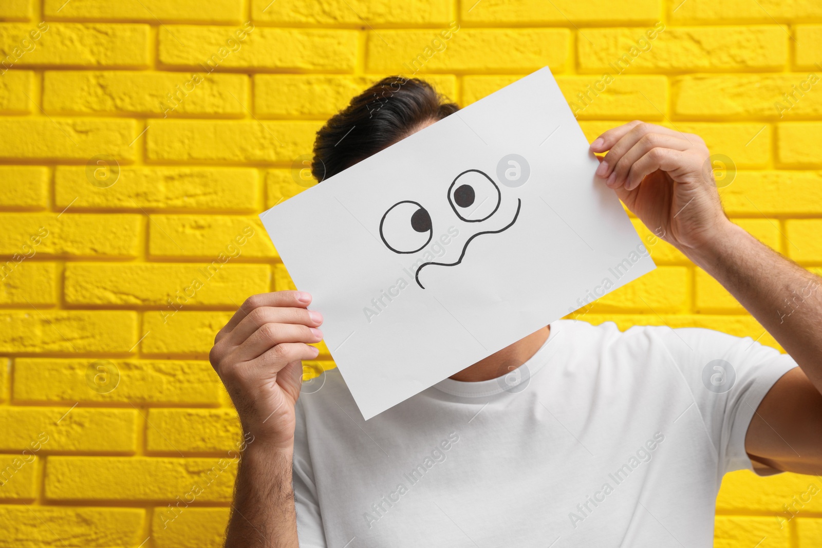 Photo of Man hiding emotions using card with drawn frowning face near yellow brick wall
