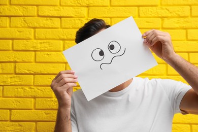 Photo of Man hiding emotions using card with drawn frowning face near yellow brick wall
