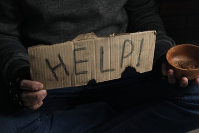 Poor homeless man with help sign holding bowl of donations on dark background, closeup