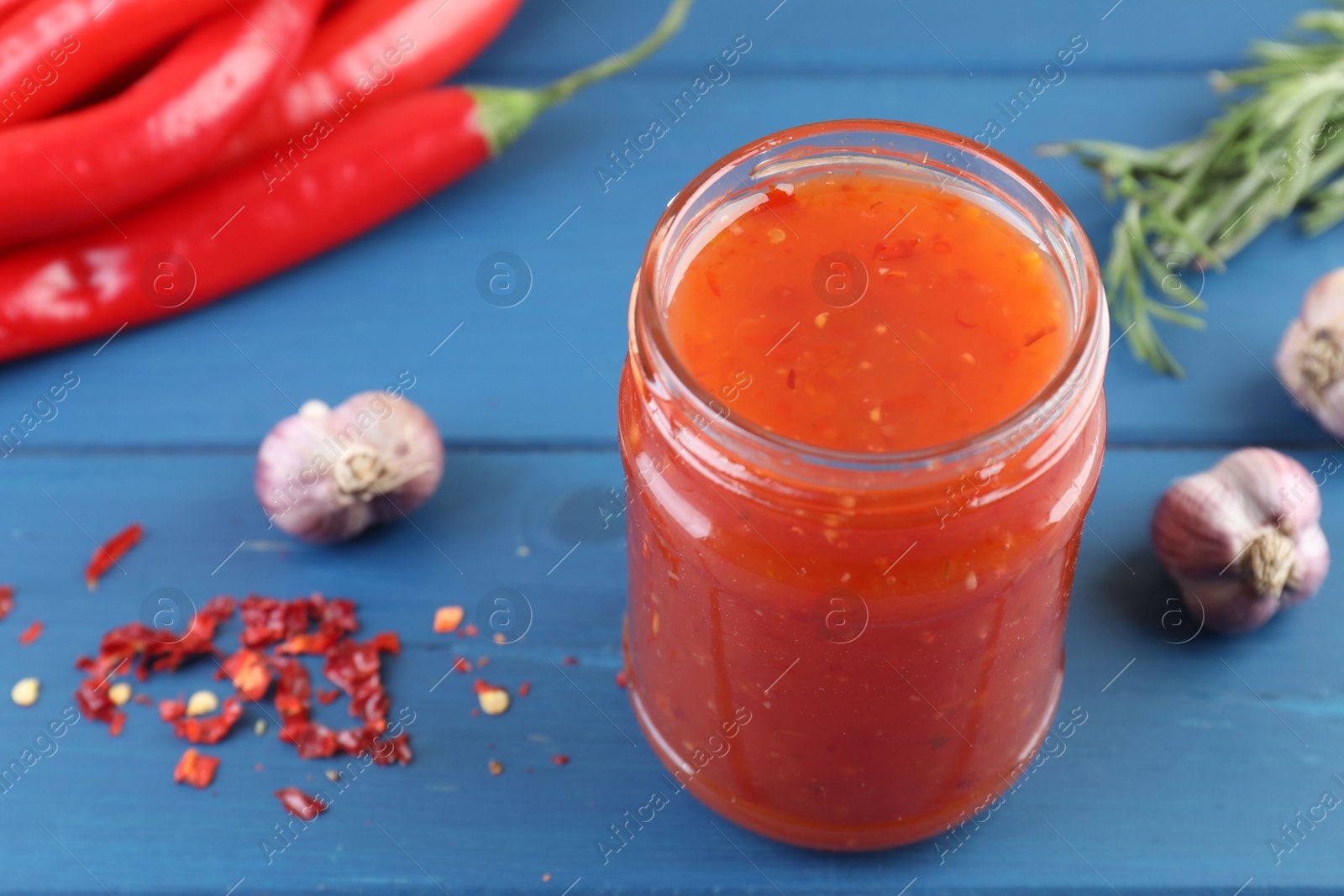 Photo of Spicy chili sauce in jar, garlic, peppers and rosemary on blue wooden table, closeup