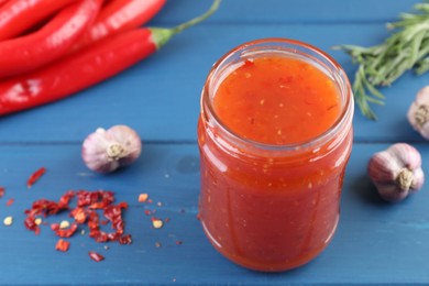 Photo of Spicy chili sauce in jar, garlic, peppers and rosemary on blue wooden table, closeup