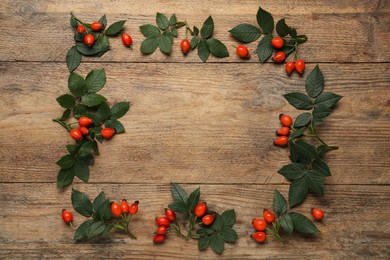 Photo of Frame of ripe rose hip berries with green leaves on wooden table, flat lay. Space for text