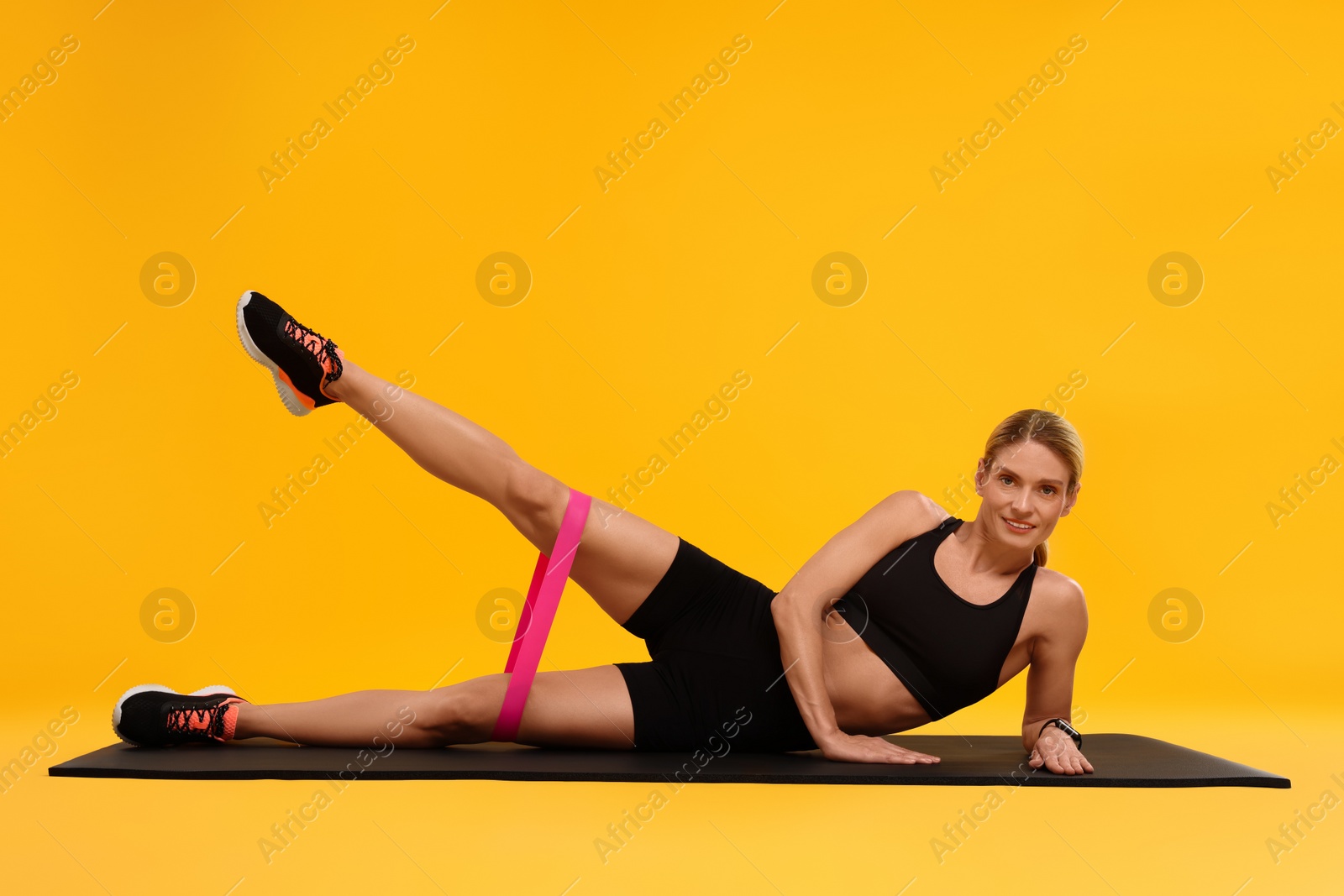Photo of Woman exercising with elastic resistance band on fitness mat against orange background
