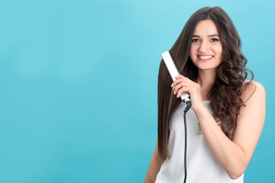 Young woman using hair iron on blue background, space for text