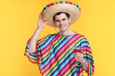 Young man in Mexican sombrero hat and poncho with cocktail on yellow background