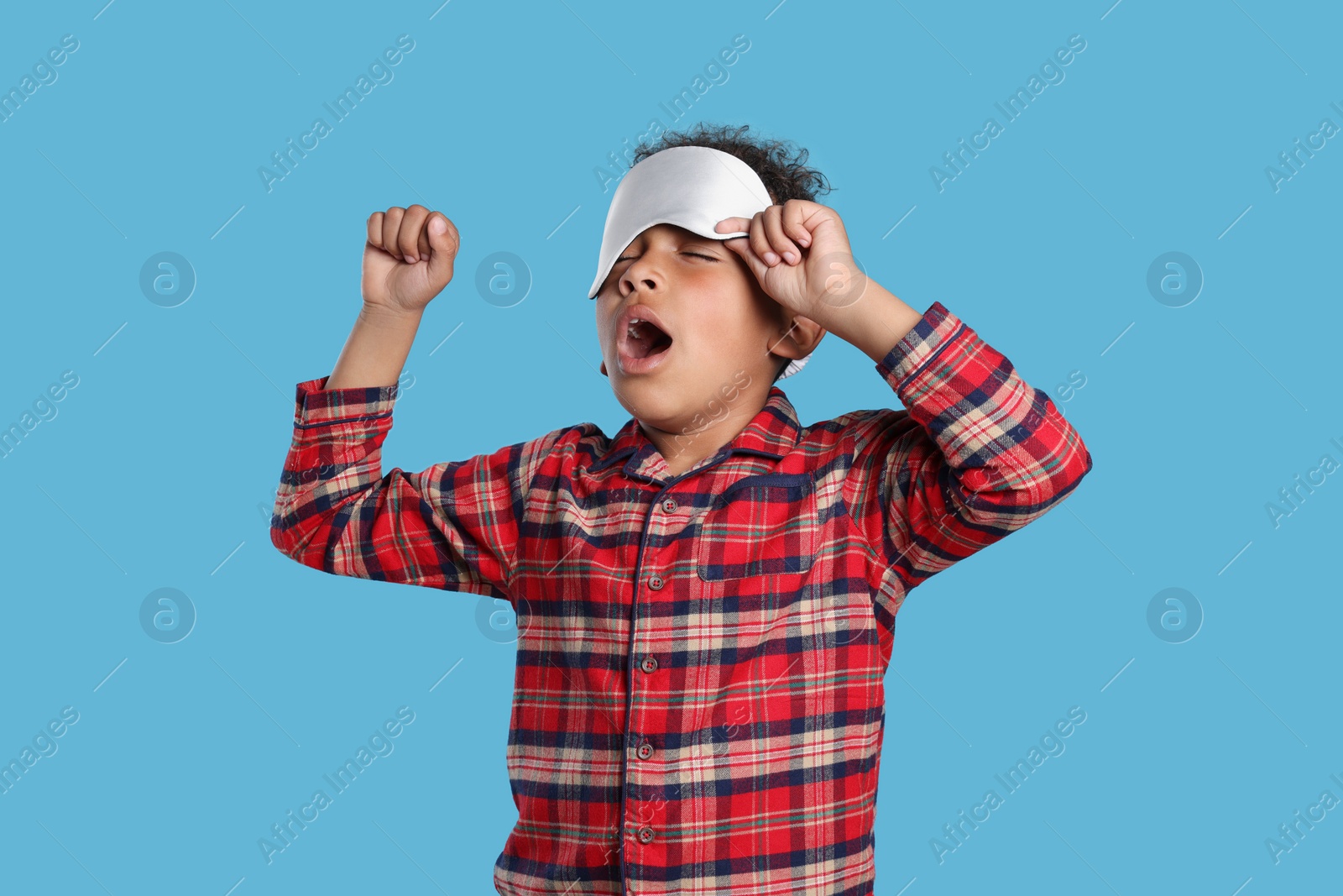 Photo of Boy with sleep mask yawning and stretching on light blue background. Insomnia problem