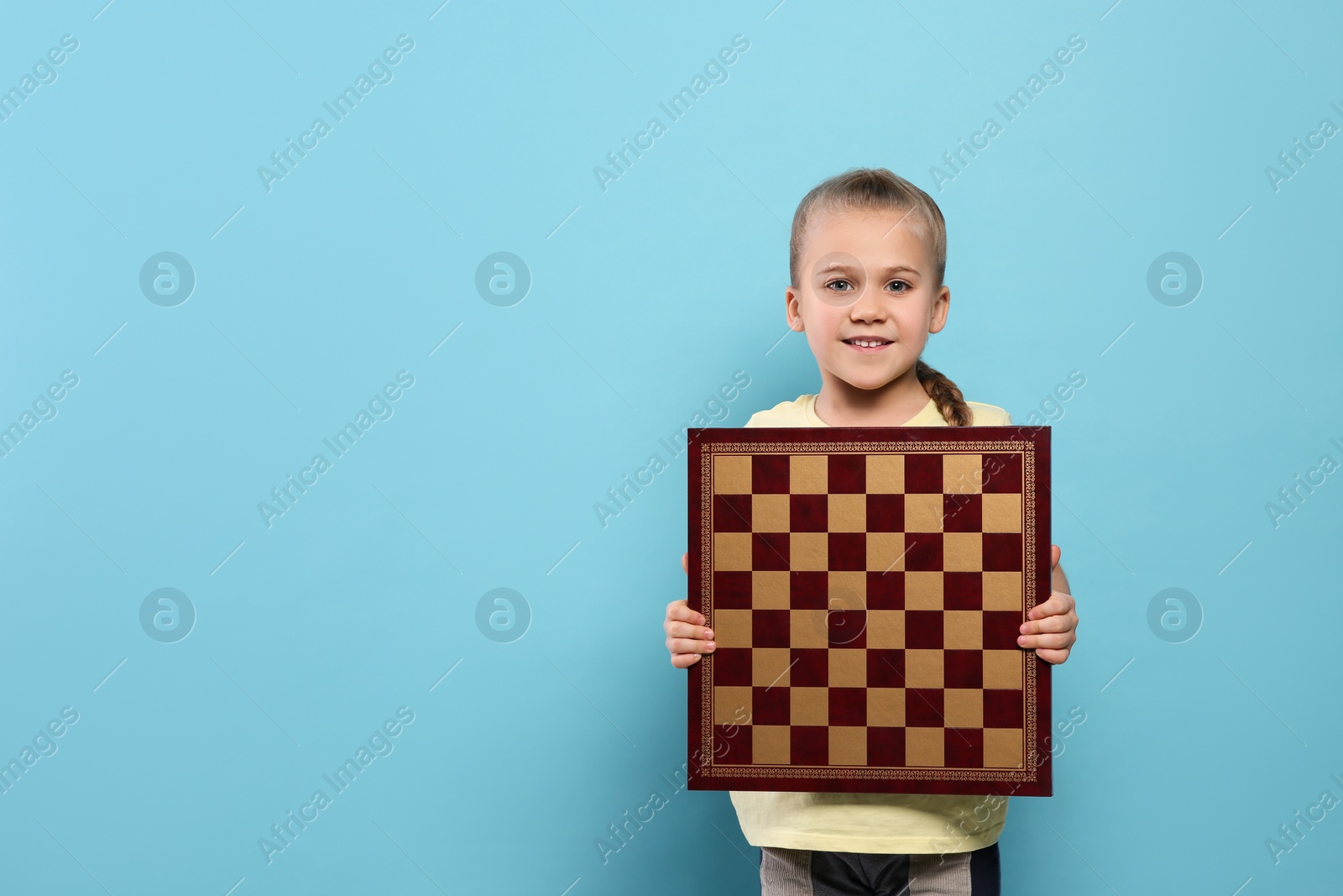 Photo of Cute girl holding chessboard on light blue background. Space for text