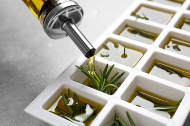 Photo of Pouring olive oil into ice cube tray with fresh rosemary at grey table, closeup