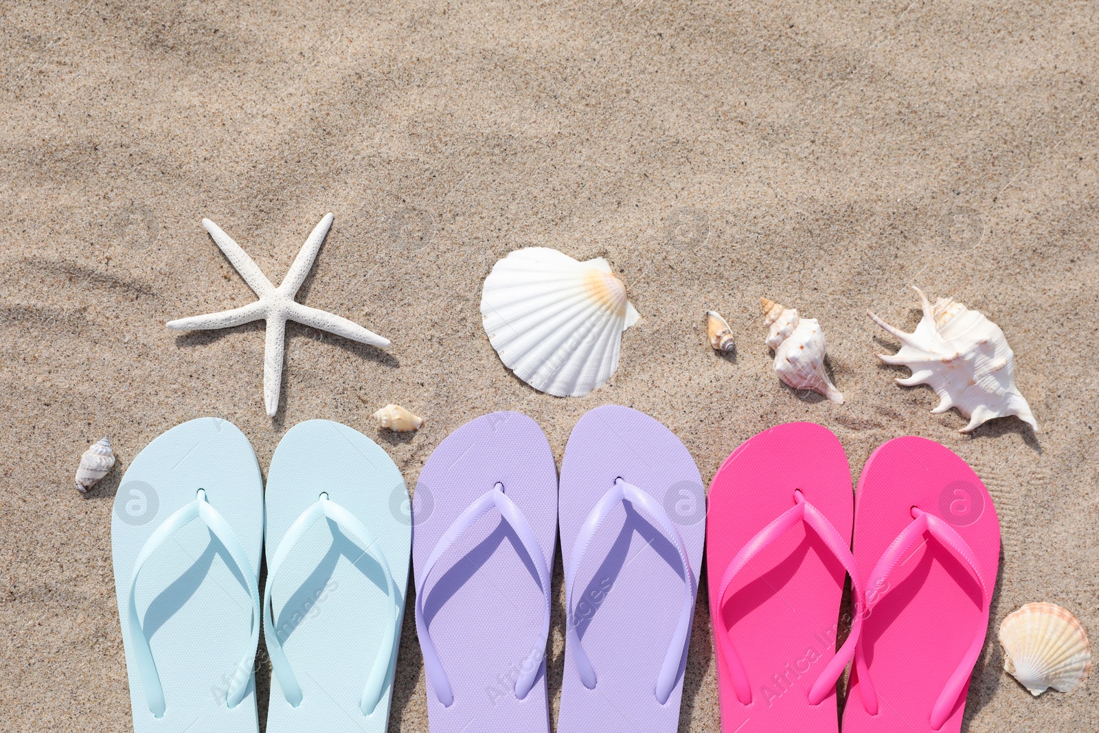 Photo of Stylish colorful flip flops, starfish and seashells on sand outdoors, flat lay. Space for text