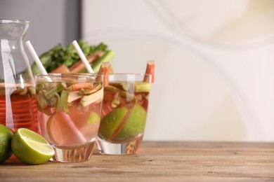Photo of Glasses and jug of tasty rhubarb cocktail with lime fruits on wooden table, space for text