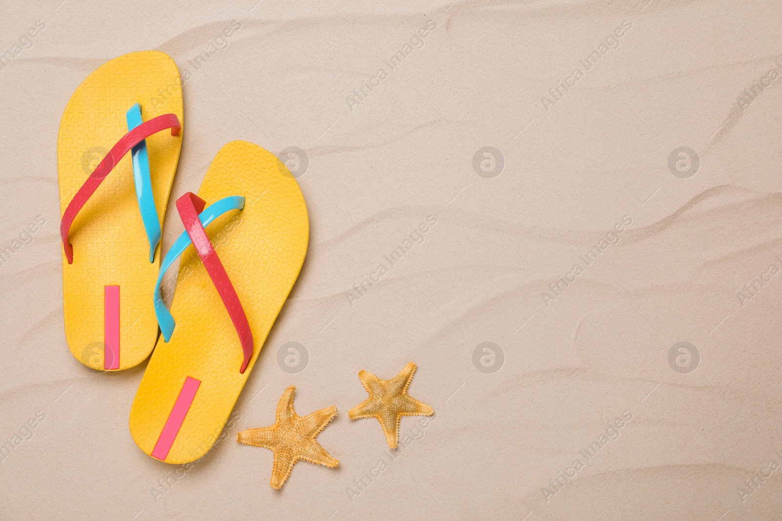 Photo of Pair of yellow flip flops and starfishes on sand, flat lay. Space for text