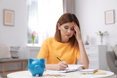 Sad woman with piggy bank and money at table indoors