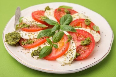 Photo of Plate of delicious Caprese salad and spoon with pesto sauce on green table, closeup