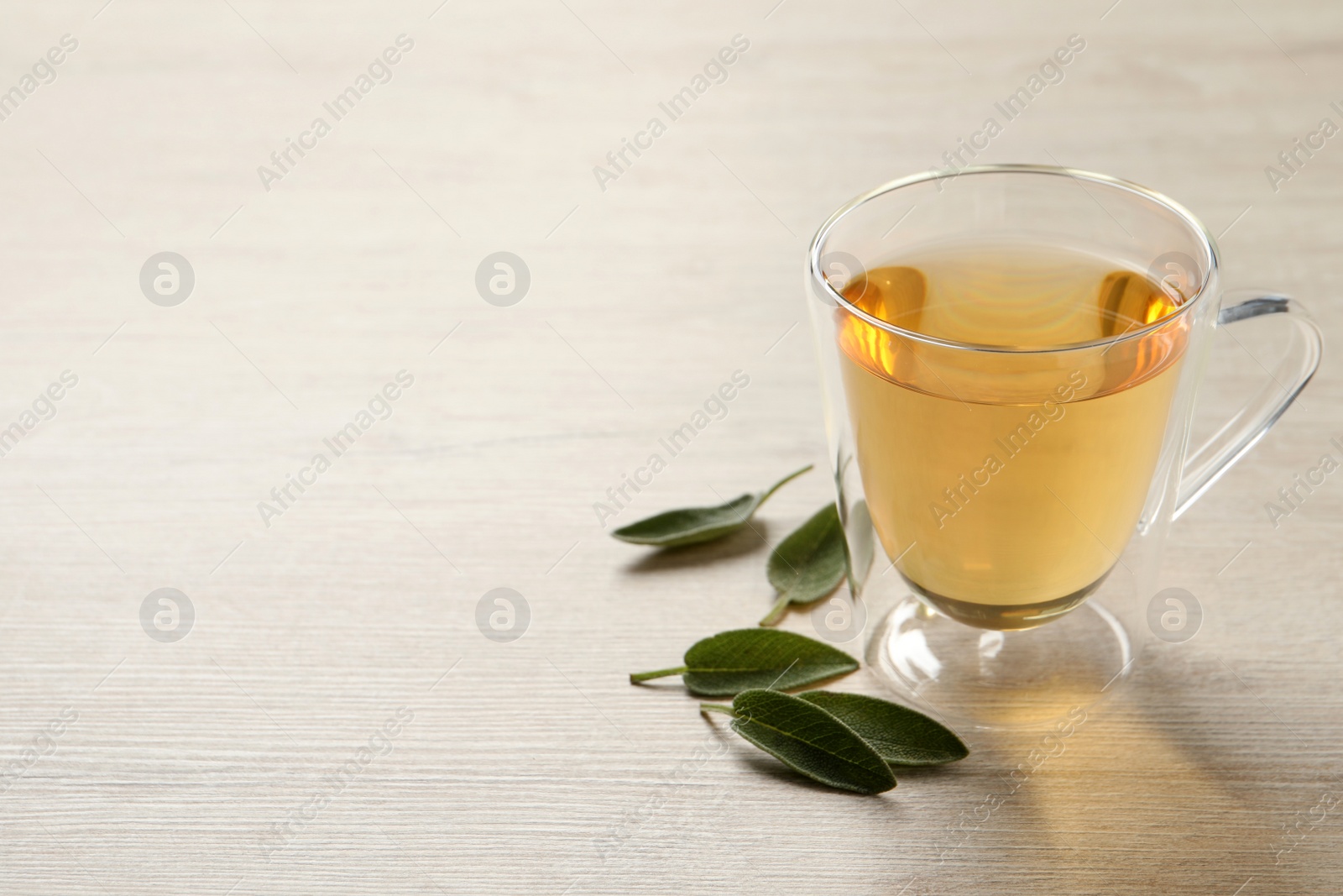 Photo of Glass cup of sage tea and green leaves on wooden table. Space for text