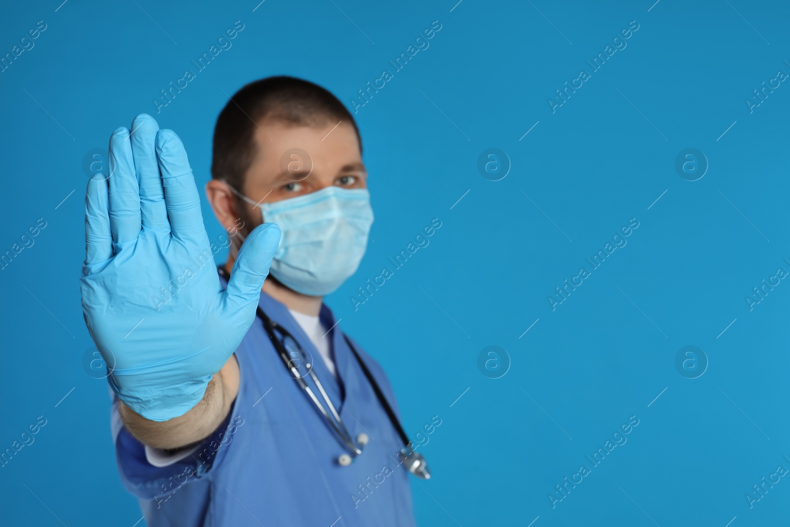 Photo of Doctor in protective mask showing stop gesture on light blue background, space for text. Prevent spreading of coronavirus