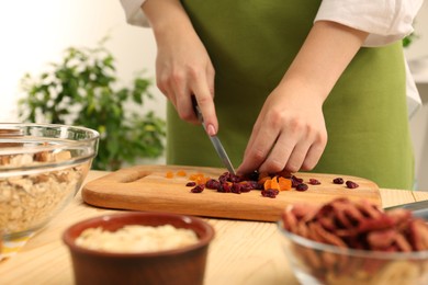 Photo of Making granola. Woman cutting dried apricots and cherries at table in kitchen, closeup