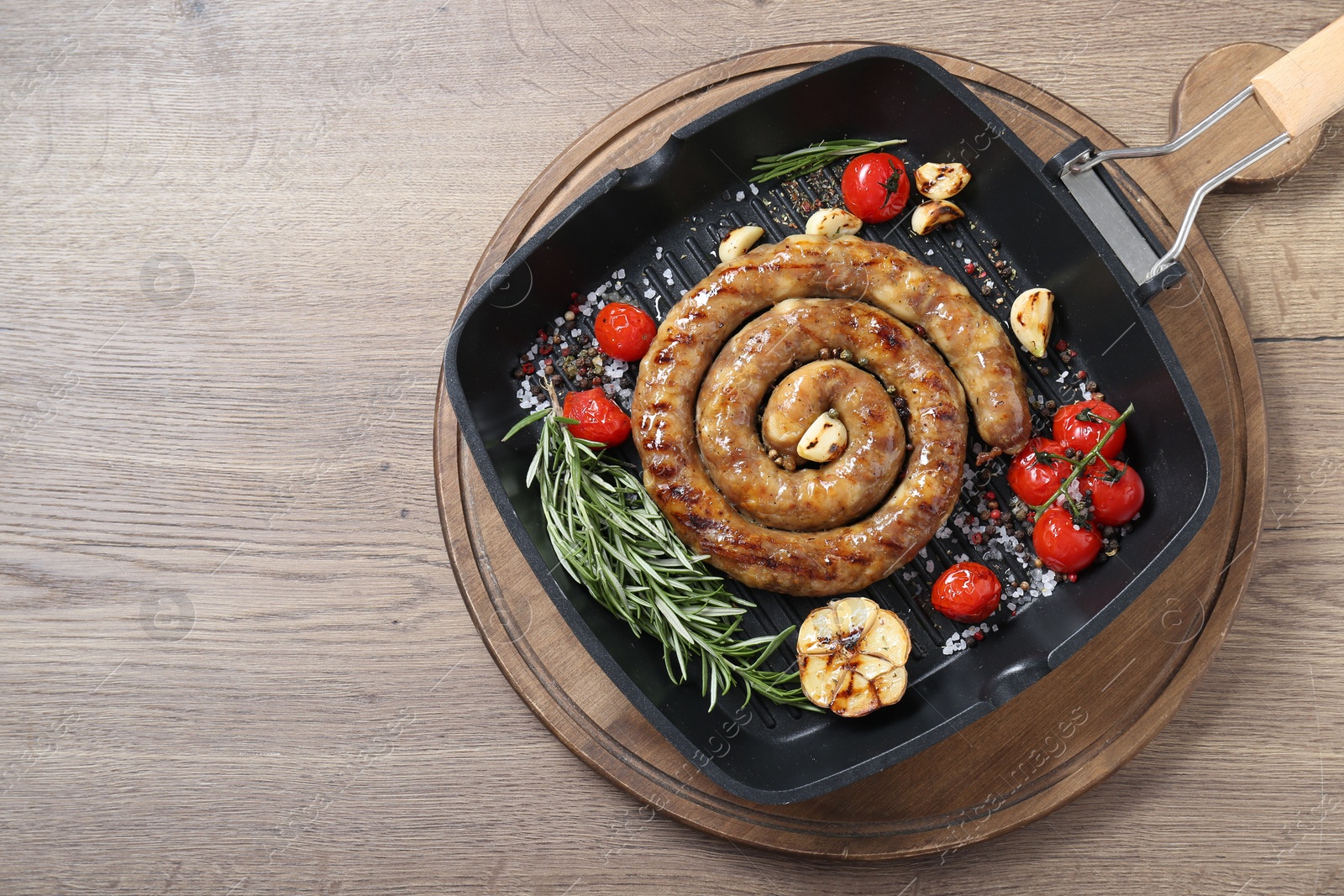 Photo of Delicious homemade sausage with garlic, tomatoes, rosemary and spices in grill pan on wooden table, top view. Space for text