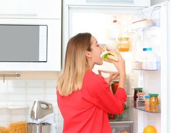 Hungry young woman eating sandwich near open refrigerator in kitchen. Failed diet