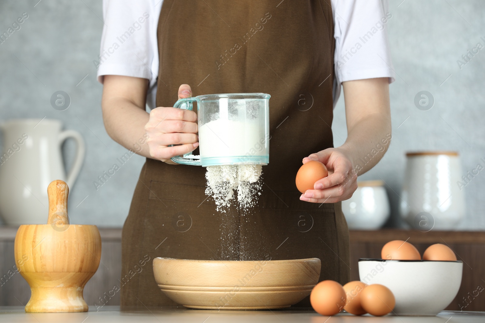 Photo of Woman sieving flour into bowl at table in kitchen, closeup