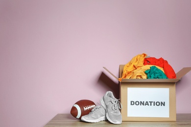 Donation box with clothes, shoes and rugby ball on table against color background. Space for text