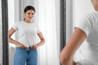 Photo of Young woman trying to put on tight jeans near mirror indoors