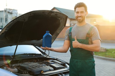 Smiling worker holding blue container of motor oil and showing thumbs up near car outdoors